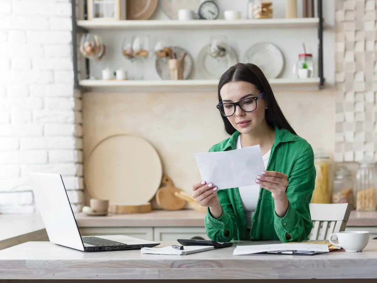 A woman stands at a kitchen counter reading a letter, an open laptop beside her.