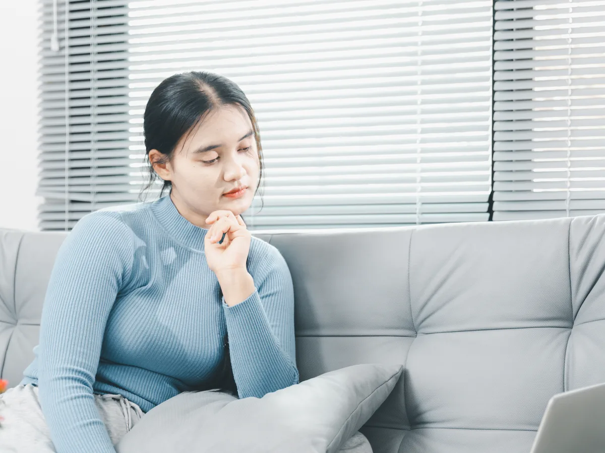 A woman sits on a sofa with a laptop, resting her chin on her hand as she reads.